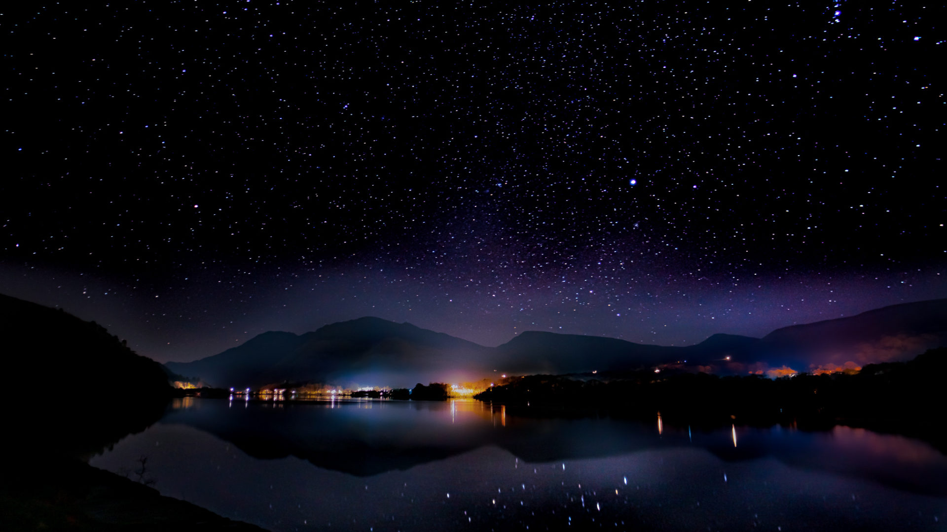 A lake at night under stars with pink and purple lights from the town on the otherside