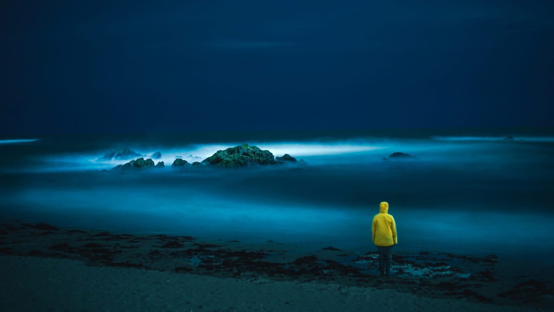 Fine art coastal photograph of dramatic Welsh sea at night with person in yellow coat on dark beach.