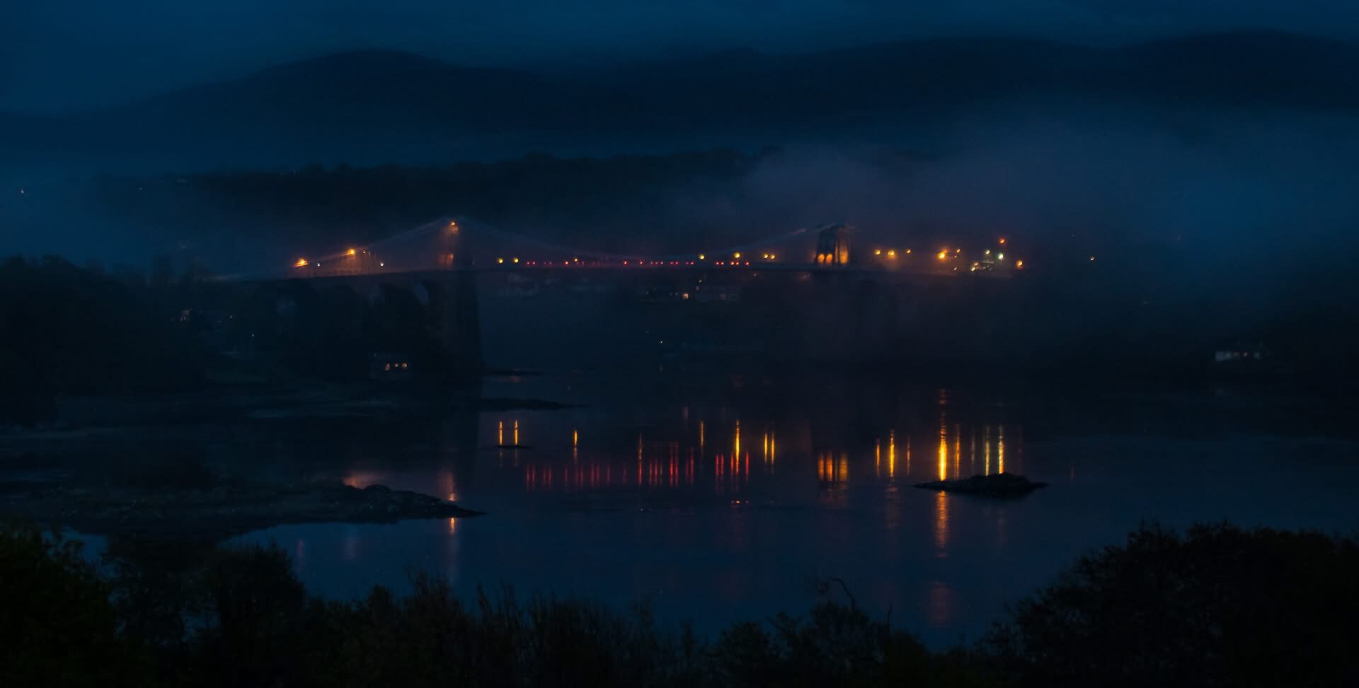 Menai bridge at night shrouded in mist. Lights from traffic cut through the foggy atmosphere.