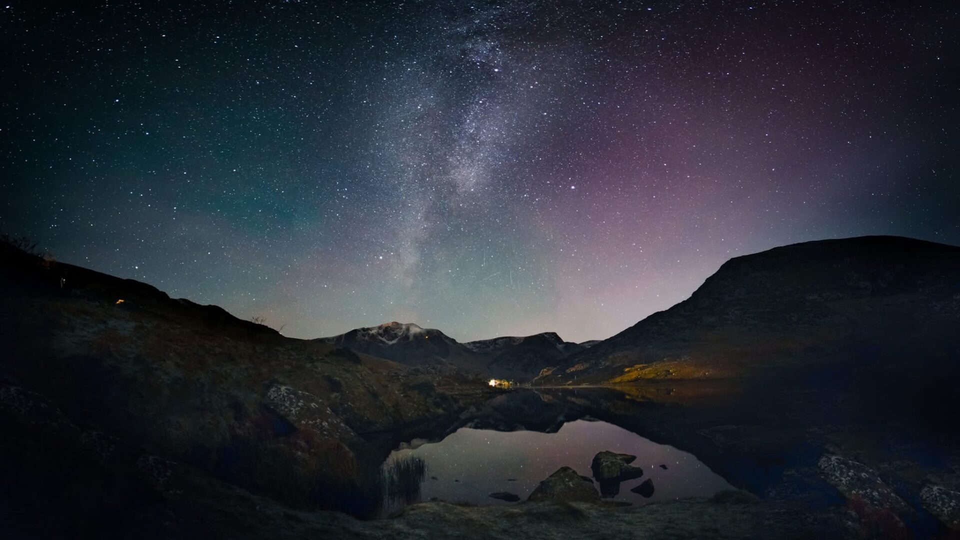 Pink Northern Lights over Llyn Ogwen in North Wales. Mountains surround the lake at night.