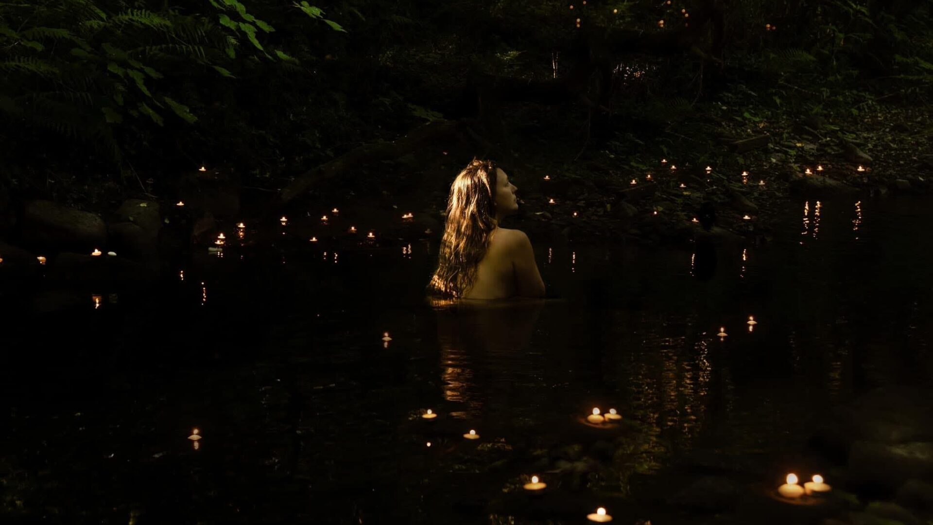 A woman with long hair surrounded by candles bathes in a pool in a wood.